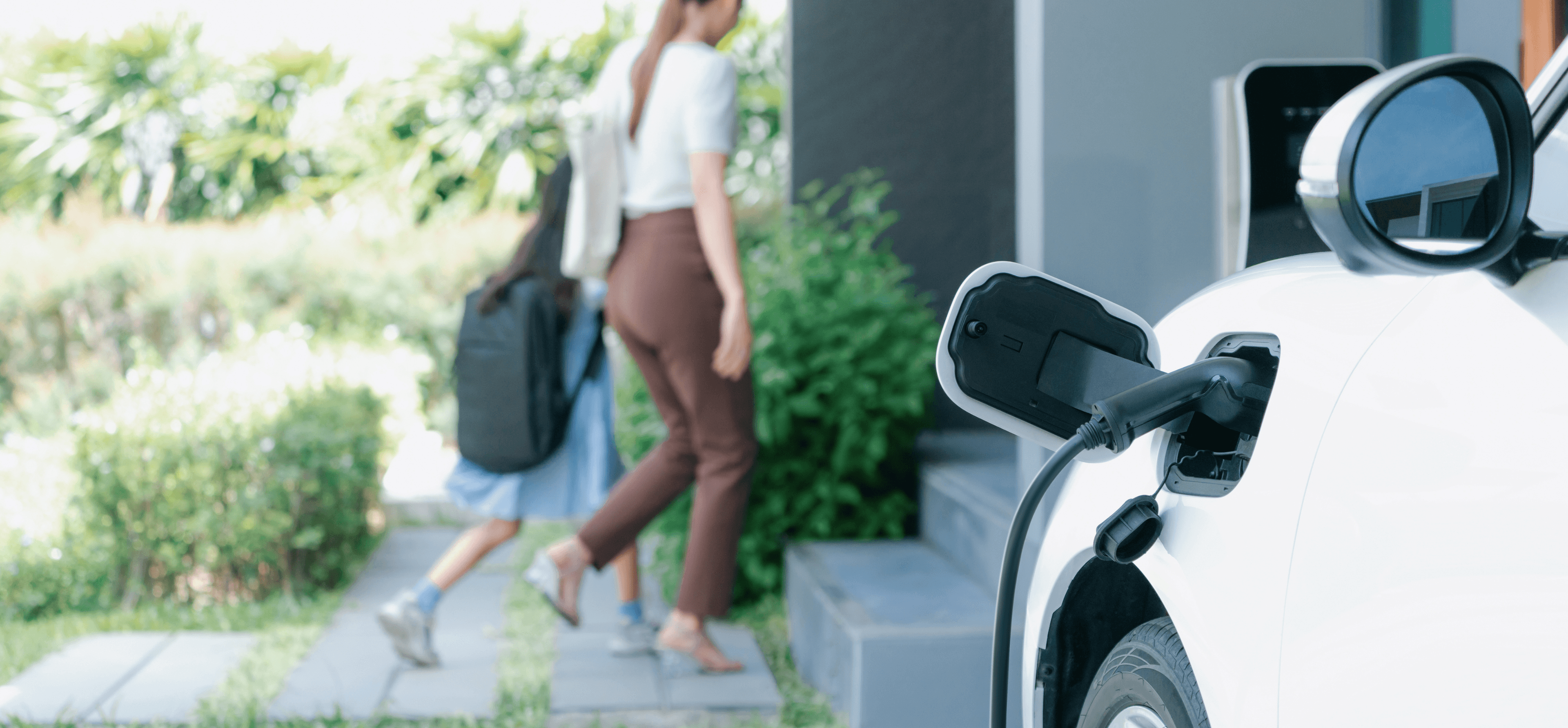 Image of white electric car plugged in and charging at home with mother and daughter walking in the background.